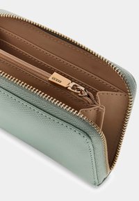 Jasnozielony portfel z fakturą i zamkiem błyskawicznym rozpinany dookoła, otwarty, ukazujący beżowe wnętrze oraz wewnętrzną kieszeń na zamek z złotym suwakiem z wygrawerowanym napisem „GUESS”.