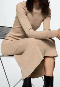 Robe longue en maille côtelée de couleur beige clair, silhouette ajustée et manches longues, assortie à des bottines noires.