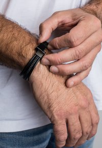 Fekete bőrből készült karkötő szövött dizájnnal, elegáns fémzárral. A csuklón világos szőr és természetes bőrszerkezet látható.