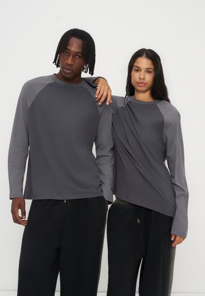 Deux personnes se tiennent côte à côte, vêtues de chemises à manches longues gris foncé assorties et de pantalons noirs, sur un fond clair uni.