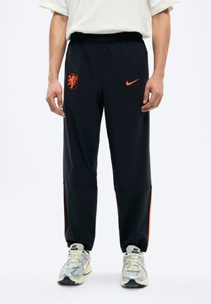 Mężczyzna ubrany w czarne spodnie dresowe Nike z pomarańczowym herbem lwa Holandii i logo Swoosh, w połączeniu z białymi butami Nike.