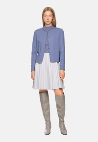 Hellblauer Cardigan mit Rundhalsausschnitt und Knopfverschluss, über einem hellgrauen, plissierten Rock getragen. Graue, kniebedeckende Stiefel runden das Outfit ab.