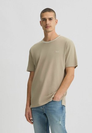 T-shirt beige a maniche corte con scollatura rotonda, caratterizzato da una sottile texture e un piccolo logo sul petto, abbinato a jeans chiari strappati.