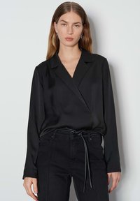 Schwarze Wickelbluse aus Satin mit langen Ärmeln und einem gepflegten Kragen, kombiniert mit hochgeschnittenen schwarzen Jeans mit einem Gürtel zum Binden.