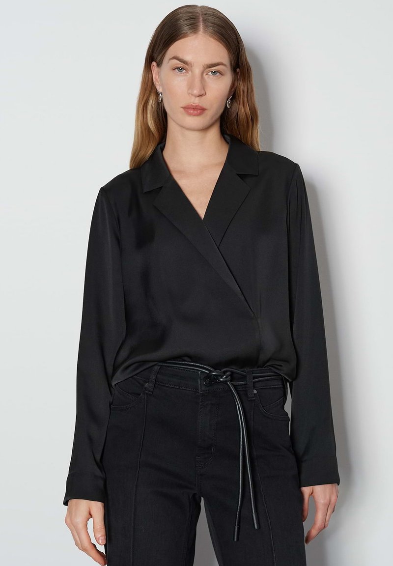 Schwarze Wickelbluse aus Satin mit langen Ärmeln und einem gepflegten Kragen, kombiniert mit hochgeschnittenen schwarzen Jeans mit einem Gürtel zum Binden.