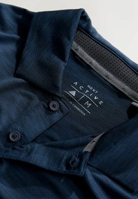 Polo bleu marine avec col, deux boutons noirs et tissu texturé. Comprend une doublure en mesh au niveau de l'encolure avec une étiquette de marque.