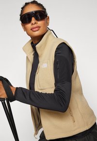 Beige fleece vest met een ritssluiting aan de voorkant, zijzakken en een logo. Draagt over een zwart langemouwen top, gecombineerd met donkere zonnebril.