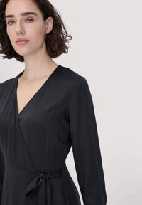 Frau mit kurzem lockigem Haar in einem schwarzen Wickelkleid mit langen Ärmeln, das in der Taille gebunden ist, vor einem schlichten hellen Hintergrund.