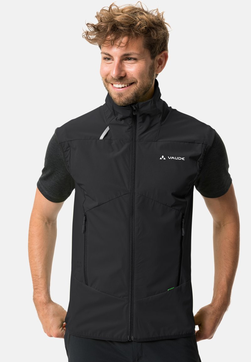 Lächelnder Mann, der eine schwarze ärmellose Jacke mit Reißverschluss über einem dunklen kurzärmeligen Hemd trägt, die Hände in die Hüften gestützt, leicht nach rechts blickend vor hellem Hintergrund.