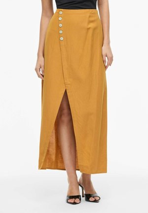 Mosterdgele midirok met een split aan de voorkant en vijf decoratieve knopen aan de linkerkant, gedragen met een zwarte top en zwarte open-teenhakken.