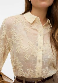 Kreemika värviga läbipaistev pluus, millel on lilleline tikandimuster, klassikaline krae ja nööpidega kinnitamine. Kerge ja tekstuuriga kangas.