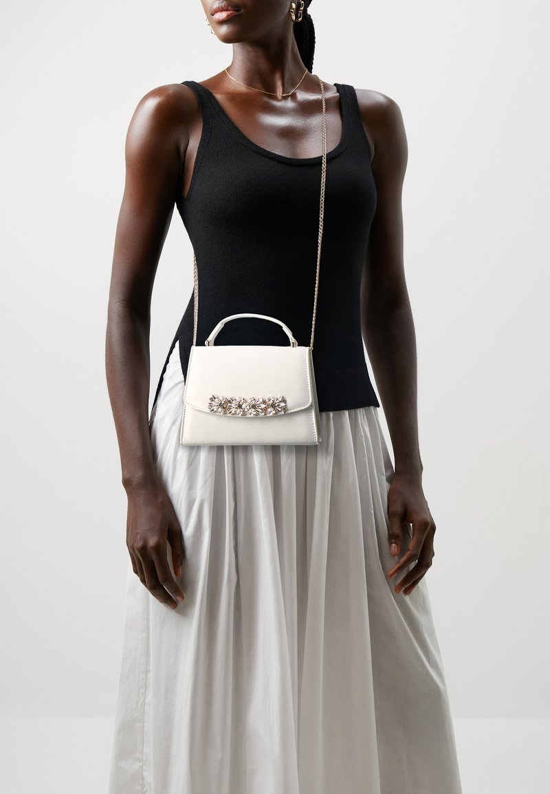 Femme portant un haut noir sans manches, une jupe plissée blanche et un petit sac à main blanc orné de fleurs sur une bandoulière chaîne.
