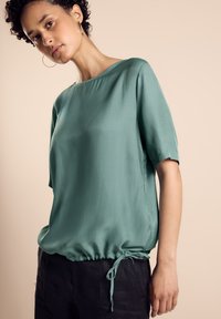 Short-sleeve top in soft, textured green fabric with a gathered drawstring hem. Smooth neckline and relaxed fit, paired with dark pants.