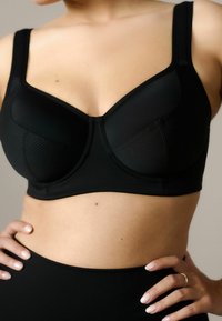 Close-up van een persoon die een zwarte ondersteunende beha met brede bandjes draagt, handen rustend op de heupen, en een natuurlijke huidskleur zichtbaar.