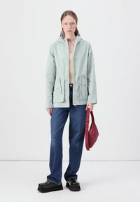 Casaco verde claro com fecho à frente, dois bolsos laterais e capuz; combinado com jeans azuis e sapatos de plataforma pretos, segurando uma bolsa vermelha.