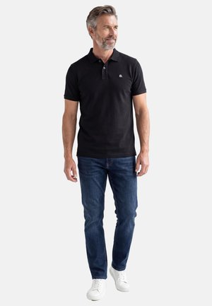 Mand med gråt hår og skæg iført en sort poloshirt, blå jeans og hvide sneakers, stående og kiggende til siden.