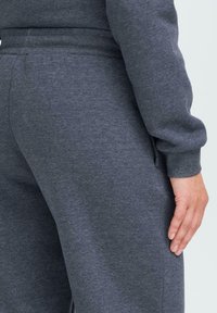 Person som har på seg mørkegrå joggebukser og matchende sweatshirt, med avslappet hånd hvilende ved siden av seg mot en lys bakgrunn.