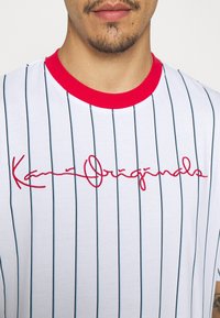 Vit kortärmad t-shirt med vertikala svarta ränder, röd krage och broderad röd text "Kaan Originals" på bröstet.