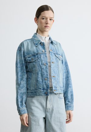 Woman with straight brown hair wearing a light blue denim jacket with faint leopard print and matching high-waisted jeans over a white zip-up top.