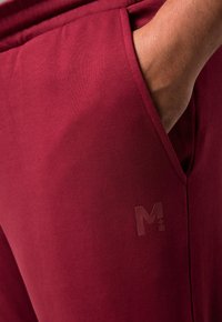 Burgunderfarbene Sporthosen mit glatter Textur, ausgestattet mit einer Seitentasche und einem subtil geprägten Logo auf der Hüfte.
