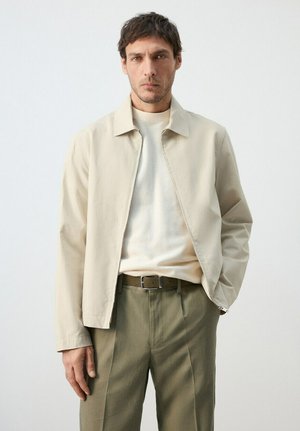 Hombre con chaqueta beige, suéter de punto claro y pantalones verde oliva, de pie con una mano en el bolsillo frente a un fondo liso.