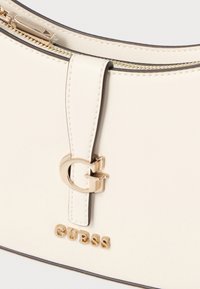 Witte leren tas met een gouden ritssluiting en hardware. Heeft een opvallend gouden "G"-logo en geperste letters "GUESS" aan de voorkant.