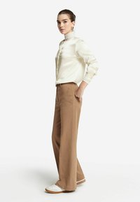 Satinblus i elfenbensfärg med hög krage och knappstängning, kombinerad med ljusbruna vida byxor och vita sneakers med bruna detaljer.