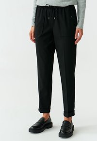Zwarte tapered broek met een elastische tailleband en trekkoord, voorzien van twee voorzakken en een gladde textuur. Gecombineerd met zwarte schoenen.