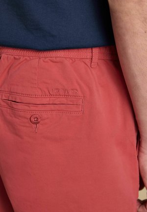 Rote Shorts mit einer geknöpften Gesäßtasche und dem darüber gestickten "Joules"-Schriftzug, getragen mit einem marineblauen Hemd, das an der Taille sichtbar ist.
