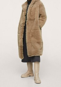 Cappotto in finta pelliccia beige con colletto a scialle, apertura frontale e tasche laterali, indossato sopra un vestito scuro e abbinato a stivali beige fino al ginocchio.