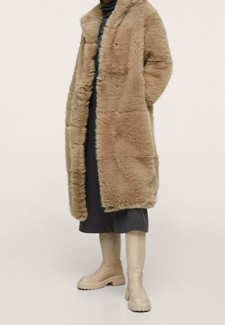 Cappotto in finta pelliccia beige con colletto a scialle, apertura frontale e tasche laterali, indossato sopra un vestito scuro e abbinato a stivali beige fino al ginocchio.