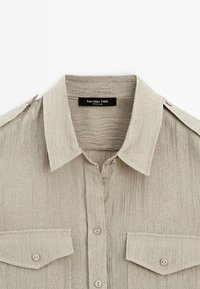 Chemise beige texturée avec col, deux poches poitrine à boutons et pattes d'épaule, étiquetée Massimo Dutti.