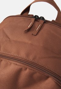 Bruine stoffen rugzak met een gestructureerde oppervlakte, voorzien van een dubbele ritssluiting en een handvat aan de bovenkant voor draaggemak.