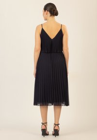 Marineblaues Faltenkleid mit V-Ausschnitt, Spaghettiträgern und fließendem Saum. Kombiniert mit schwarzen, geschnittenen High Heels.
