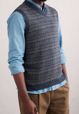 Mężczyzna ubrany w niebieską, wzorzystą dzianinową kamizelkę na jasnoniebieskiej, podwiniętej koszuli oraz brązowe spodnie na jednolitym tle.