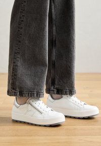 Vit sneaker med en texturerad gummisula och metalldragkedjedetalj. Slät läderöverdel och kontrasterande grå detaljer. Bärs med grå jeans.