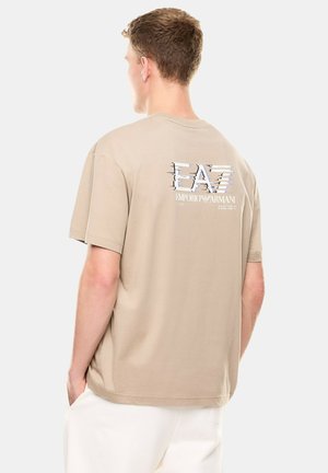 Giovane uomo che indossa una T-shirt beige a maniche corte con il logo EA7 Emporio Armani sul retro, abbinata a pantaloni bianchi, in piedi di lato.