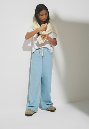 Jeune femme debout contre un mur blanc, portant un t-shirt blanc, un jean large bleu clair, des chaussures beiges, ajustant un pull crème drapé sur ses épaules.