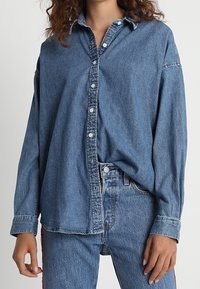 Denimskjorta med knappar, medelblå, lös passform, klassisk krage, har vita knappar, långa ärmar och subtil textur. Bärs över jeans.