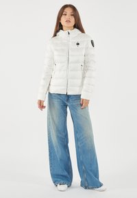 Giacca a vento bianca con design trapuntato orizzontale, zip anteriore e cappuccio. Abbinata a jeans a gamba larga azzurri e scarpe bianche.
