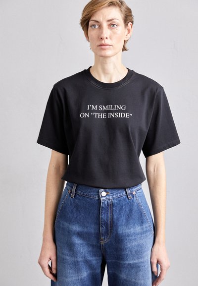 Tricou din bumbac negru cu mâneci scurte, având text alb: "ZÂMBESC ÎN INTERIOR". Purtați cu blugi albaștri cu talie înaltă.