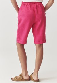 Pink linen shorts with an elastic waistband, side pockets, and rolled cuffs. Worn with brown slip-on sandals.