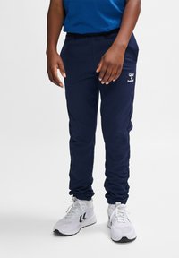 Marine blauwe joggingbroek met een elastische tailleband en gebundelde zoom, gemaakt van zachte stof; heeft een wit logo op de bovenbeen. Draagt met witte sportschoenen.
