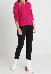 Donna che indossa un maglione testurizzato di un rosa acceso, jeans neri slim e stivaletti bianchi con tacco a blocco rosso, in piedi su uno sfondo neutro.