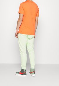 Muž v oranžovém tričku s krátkým rukávem, světle zelených běžeckých kalhotách a zeleno-červených teniskách Nike s vysokým střihém, stojící na čistém bílém pozadí.