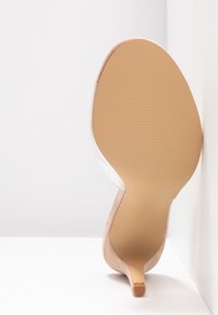 Chaussure à talons aiguilles nude avec une semelle lisse couleur tan, ornée d'un bout pointu et d'un accent en matériau transparent sur les côtés, le tout présenté sur un fond blanc.