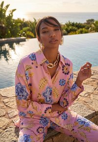 Žena oblečená v ružovej kvetinovej košeli a nohaviciach, s veľkým zlatým náhrdelníkom a náušnicami, sediaca pri bazéne s zeleňou v pozadí.