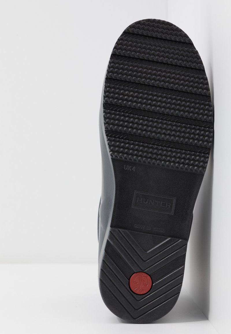 La suela de la bota de goma negra presenta una superficie texturizada con un acento circular rojo, el nombre de la marca en relieve y un patrón de tracción distintivo.