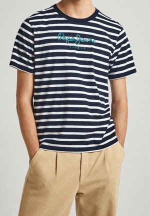 Homme portant un t-shirt rayé bleu marine et blanc Pepe Jeans London avec un pantalon beige et les mains dans les poches, sur un fond uni.