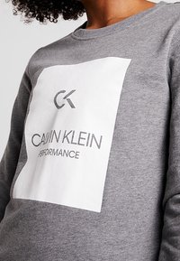 Sweat-shirt gris avec un logo rectangulaire blanc affichant "CK" et "CALVIN KLEIN PERFORMANCE" en texte noir. Encolure ronde et poignets côtelés.